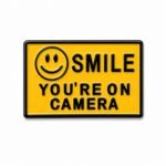 ستيكر معدني أصفر بتصميم Smile You're on Camera مع أيقونة الوجه المبتسم.